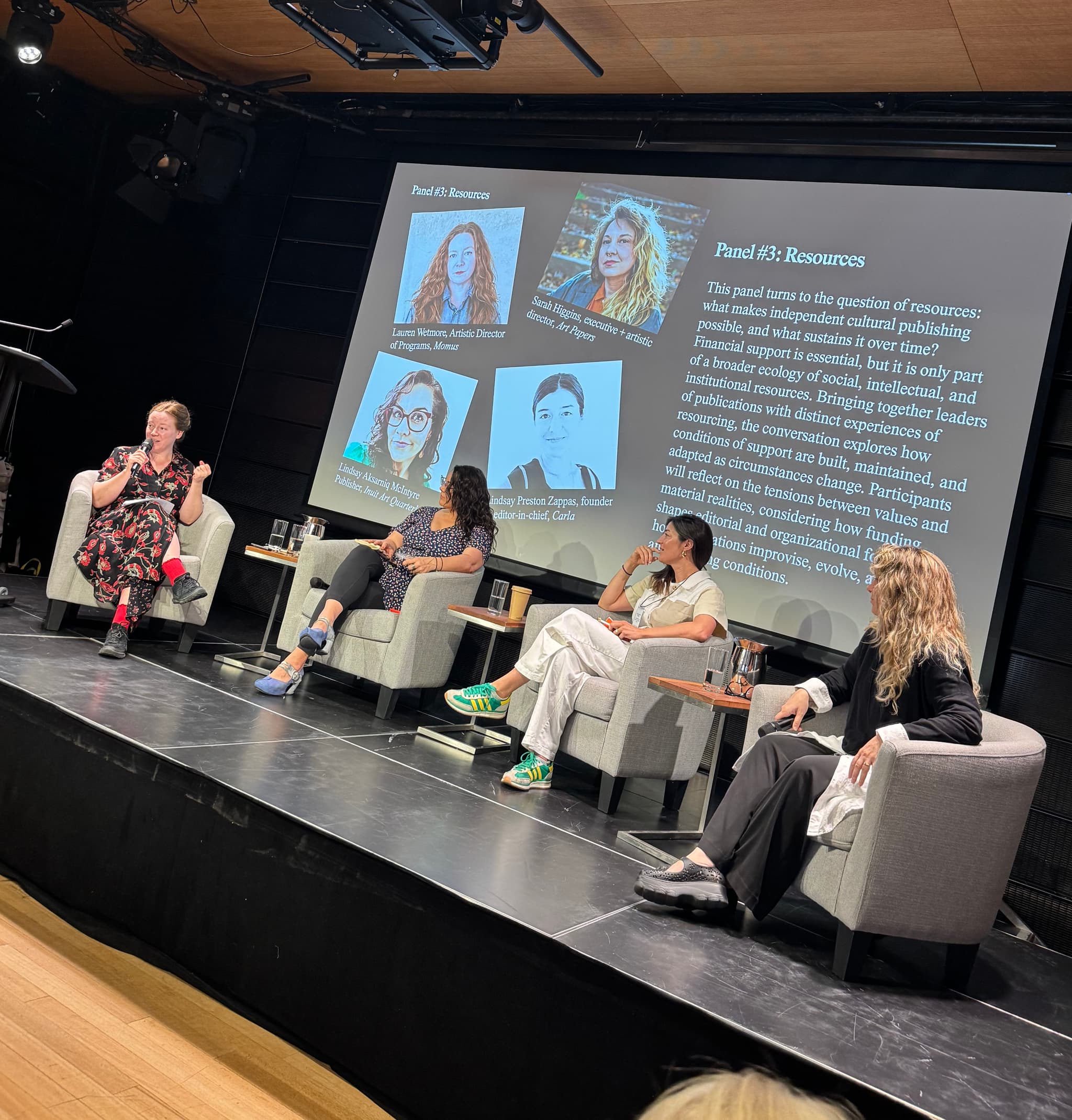 Four panelists converse, seated in front of a screen on a stage.