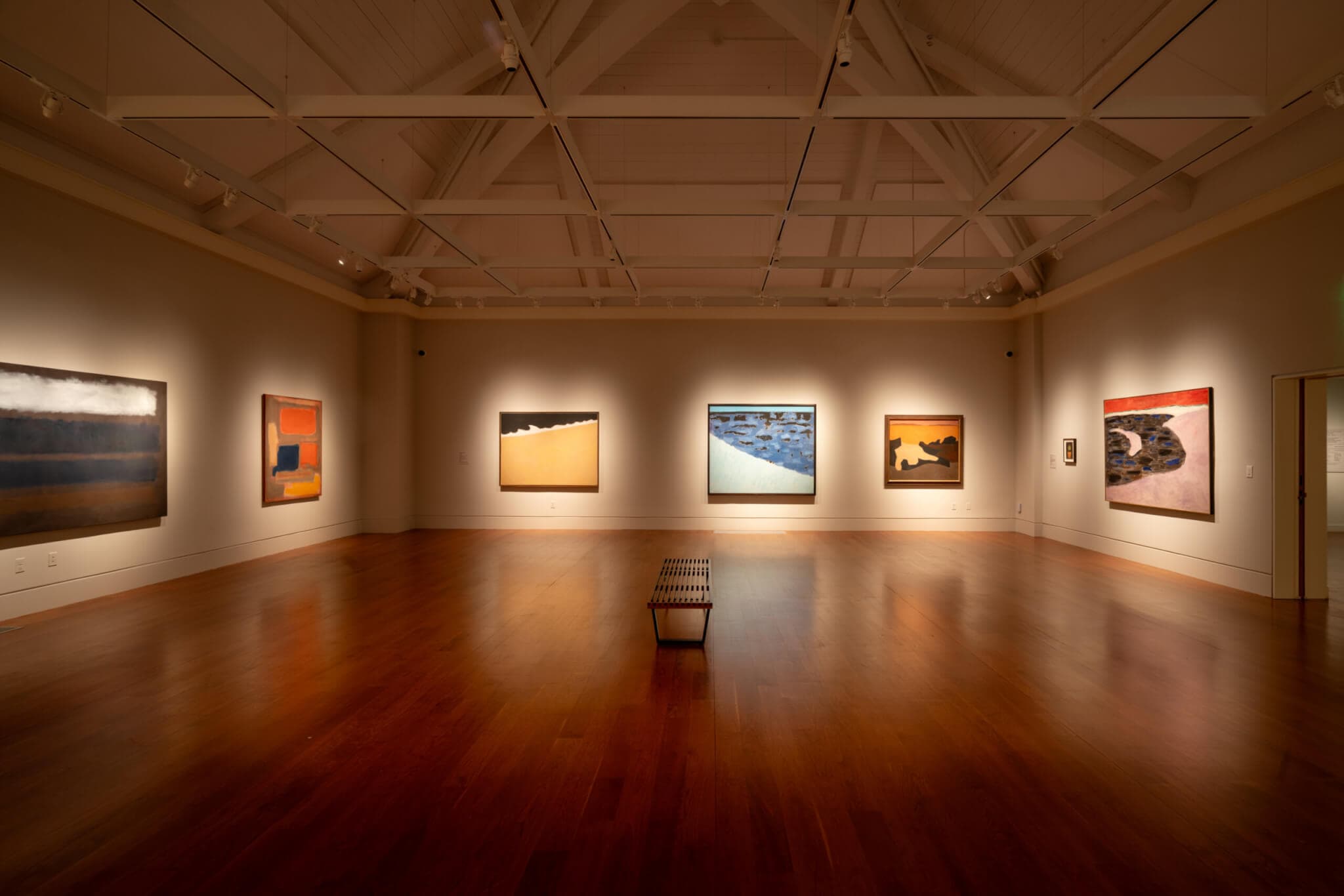 Large paintings lit from above line the walls of a darkened gallery space.