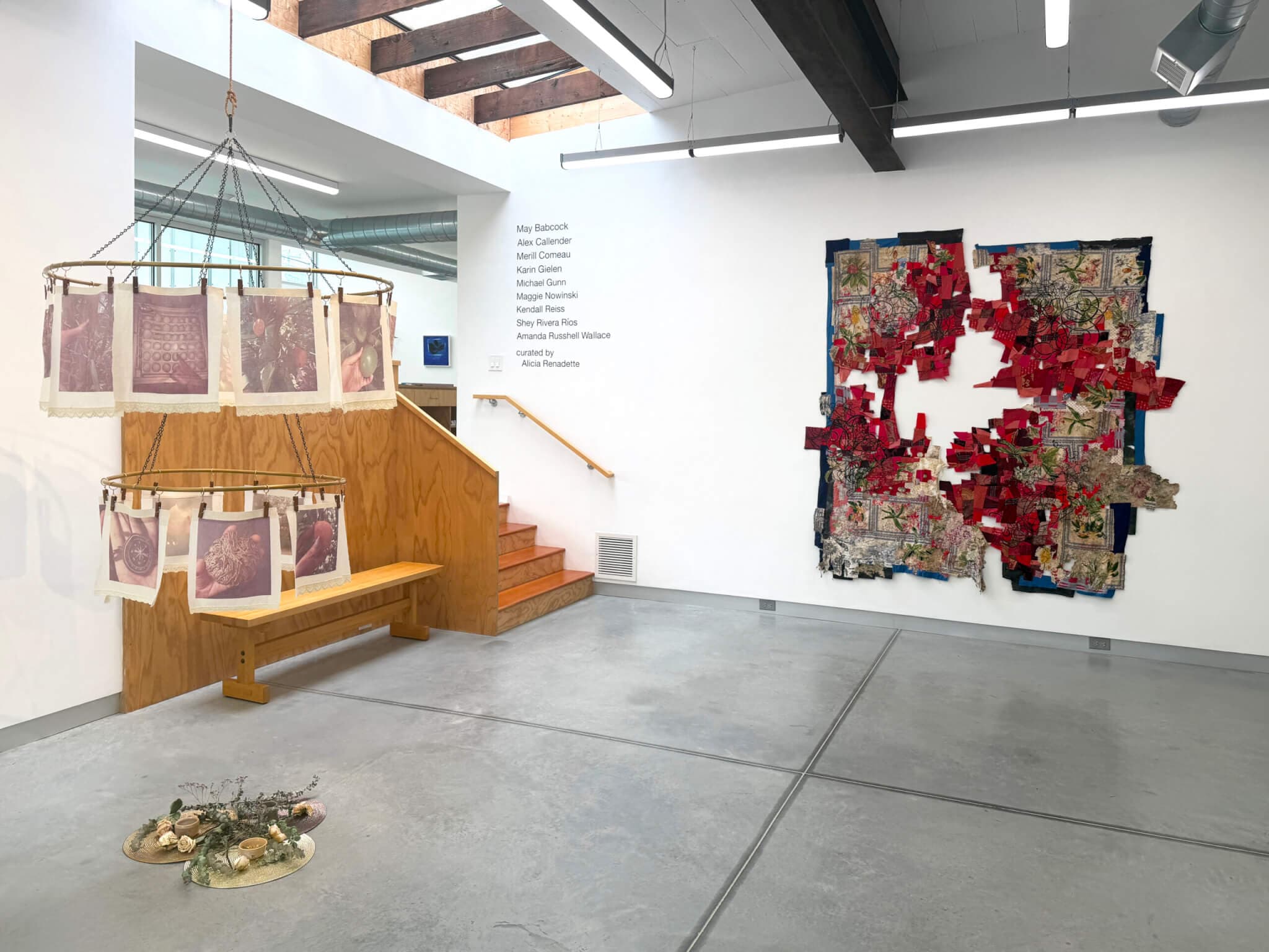 A two-tier sculpture hangs to the left of a large, textile collage on a gallery wall.