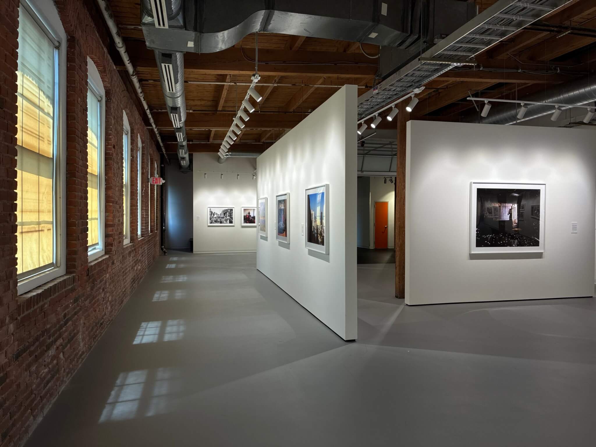 A gallery with an exposed brick wall lined with shaded windows, featuring color and black-and-white-photographs mounted on freestanding walls.